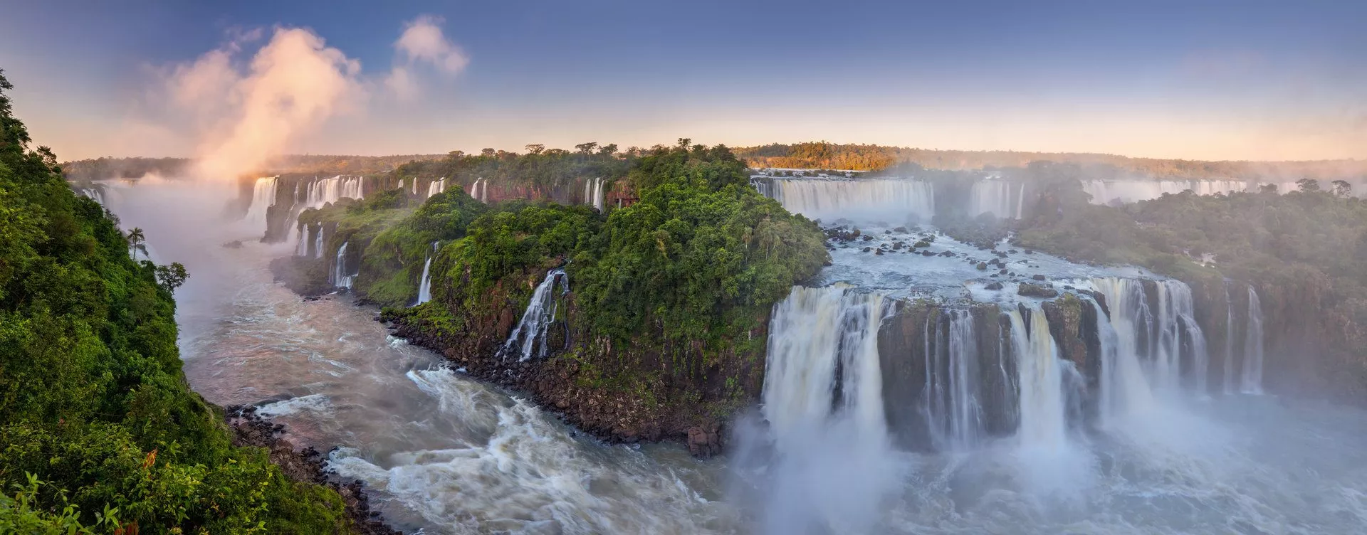 Panoramablick auf die Iguazú-Wasserfälle, umgeben von üppiger Vegetation und aufsteigenden Nebelschwaden.