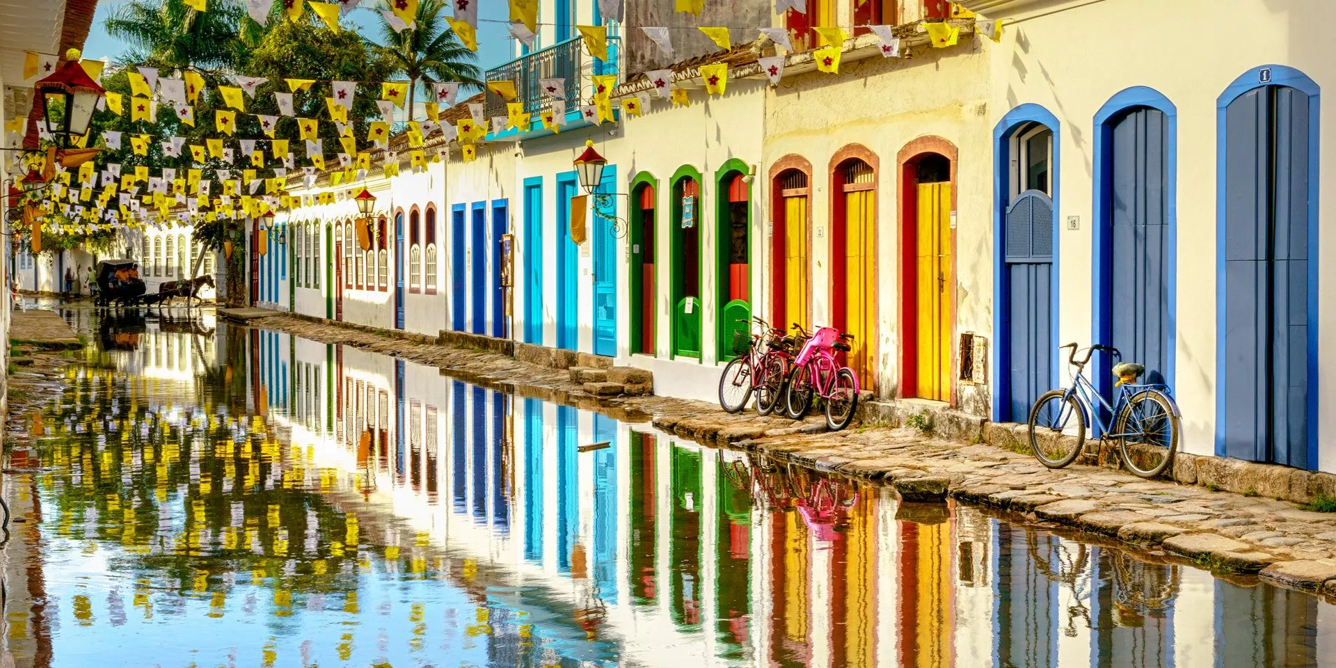 Bunte koloniale Gebäude reflektieren sich in einem Wasserspiegel auf der Straße von Paraty, Brasilien, mit Fahrrädern und einem Pferdegespann im Hintergrund.