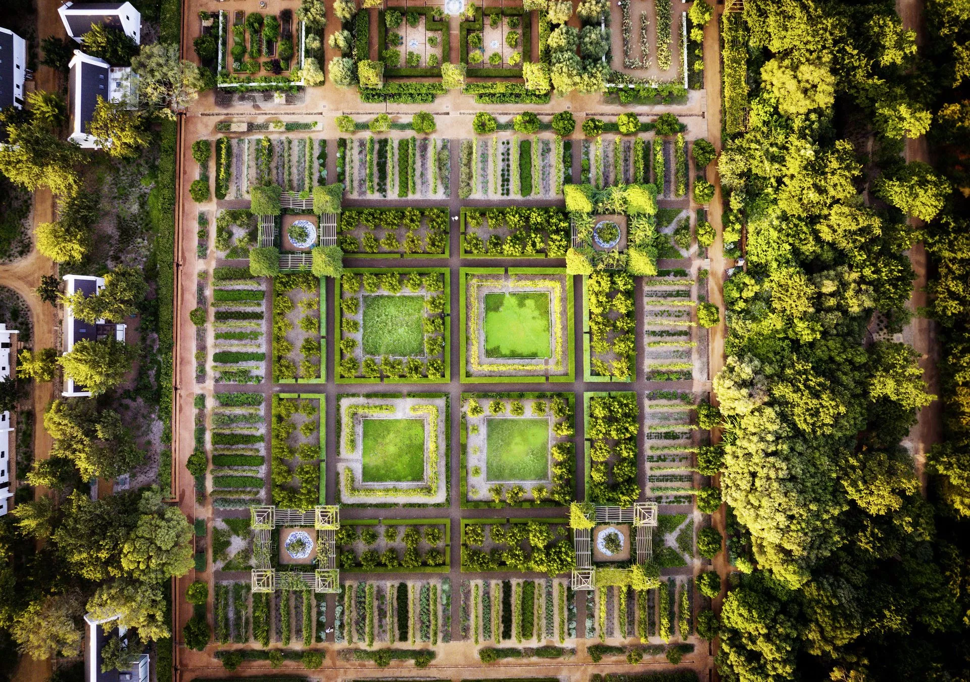 Luftaufnahme eines gepflegten Gartens mit symmetrischen Beeten, grünen Flächen und umgebenden Bäumen, sowie angrenzenden Wegen und Gebäuden.